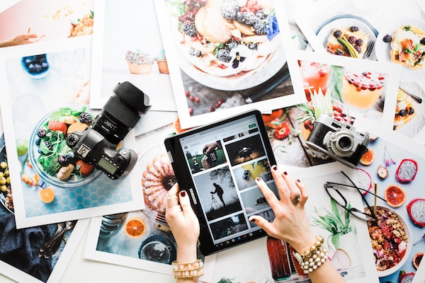 Hands holding a tablet with photos pulled up, photos spread across the table, photos of food, nice camera sitting on top of photos on table
